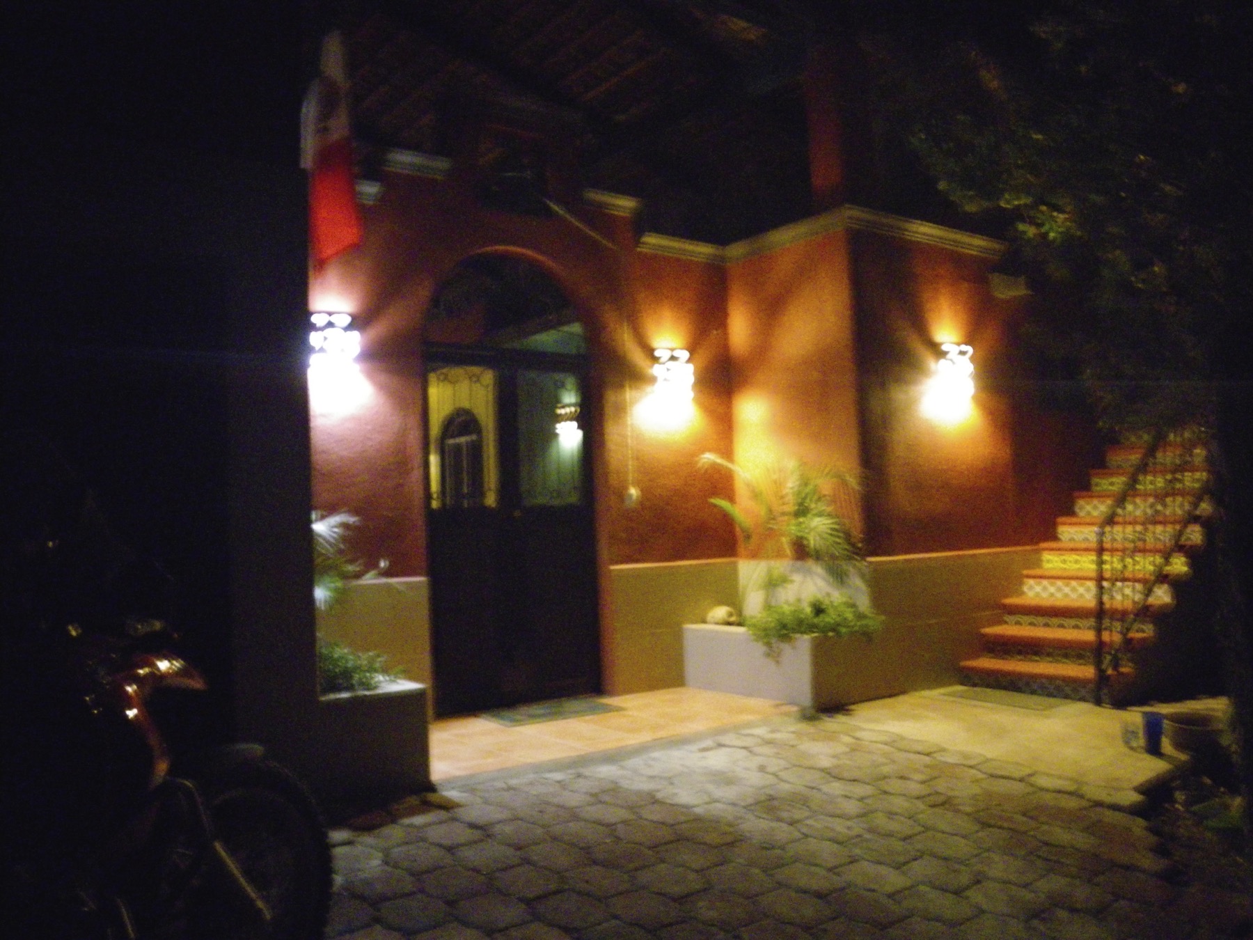 Red stucco entryway at night, lit by wall sconces, with a tiled staircase and a Mexican flag above the door