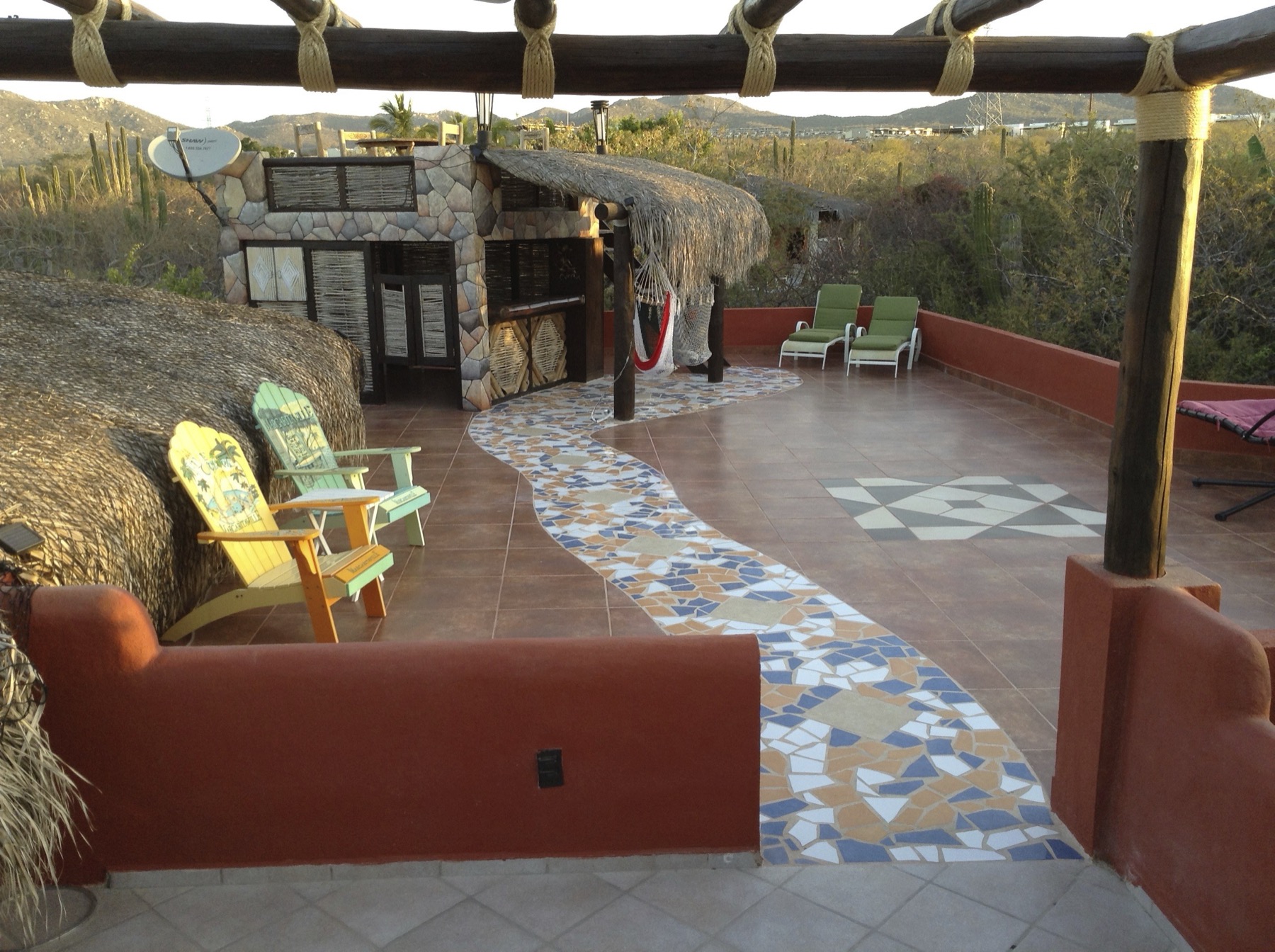 Rooftop terrace with a mosaic tile pathway, a stone palapa-roofed bar, a hammock, and lounge chairs facing the mountains